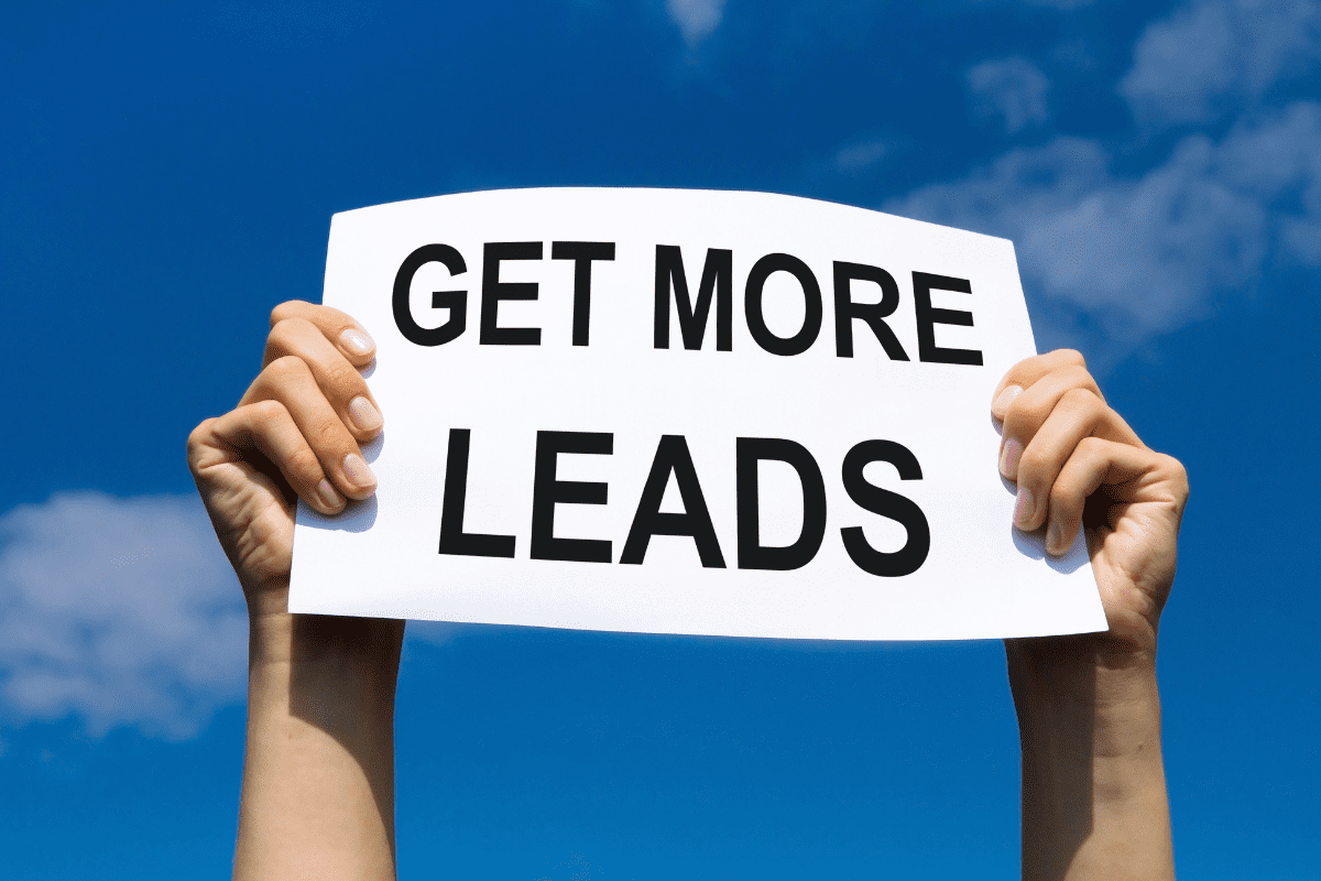 Two hands holding up a white sign with bold black text that reads “GET MORE LEADS” against a bright blue sky with scattered clouds.