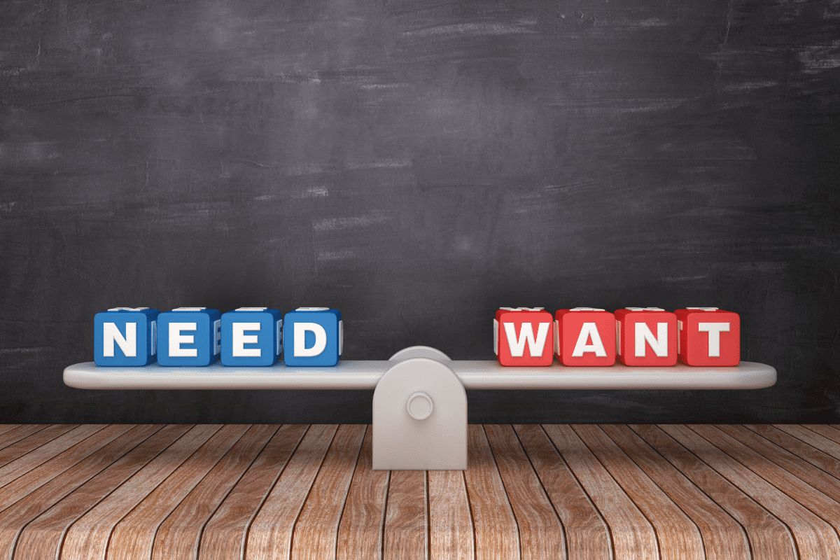 A balancing scale on a wooden surface with blue lettered cubes spelling “NEED” on the left side and red cubes spelling “WANT” on the right side, set against a chalkboard background—illustrating the contrast between needs and wants.